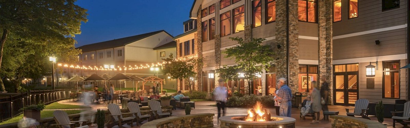 An evening outdoor scene features people around a fire pit with string lights and Adirondack chairs, against a backdrop of a large, lit building.