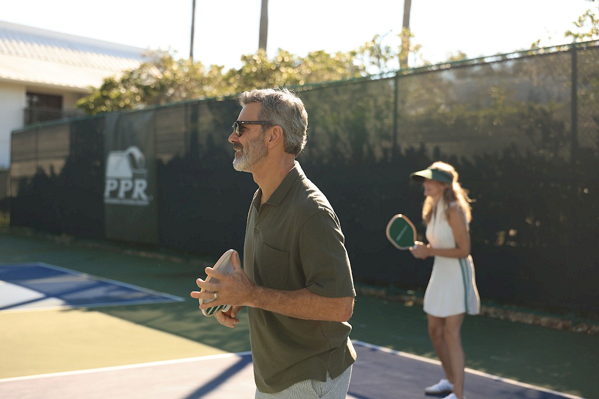 A man in a green shirt tossing a tennis ball on a court, with a woman in white in the background holding a racket near a fence. end.