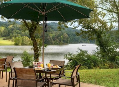 Outdoor dining area with a green umbrella, table, and chairs set near a scenic lake surrounded by trees and hills.
