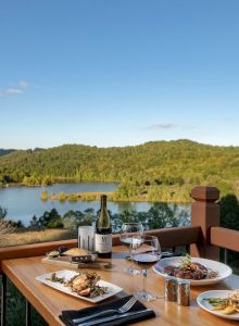 A table set with dishes of food, a bottle of wine, and glasses overlooking a scenic lake and forested hills under a clear sky.