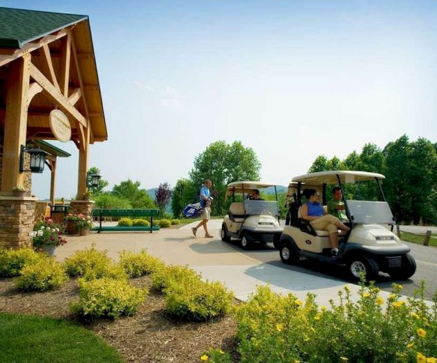 People at a golf course with two golf carts and a landscaped area, one person carrying golf clubs, near a building with a wooden structure.
