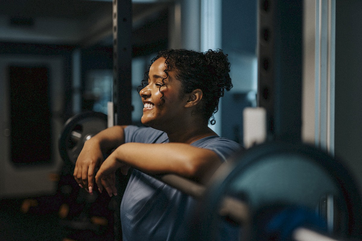 A person in a gym leans on a barbell, smiling, with a blurred background indicating equipment and dim lighting in the scene.