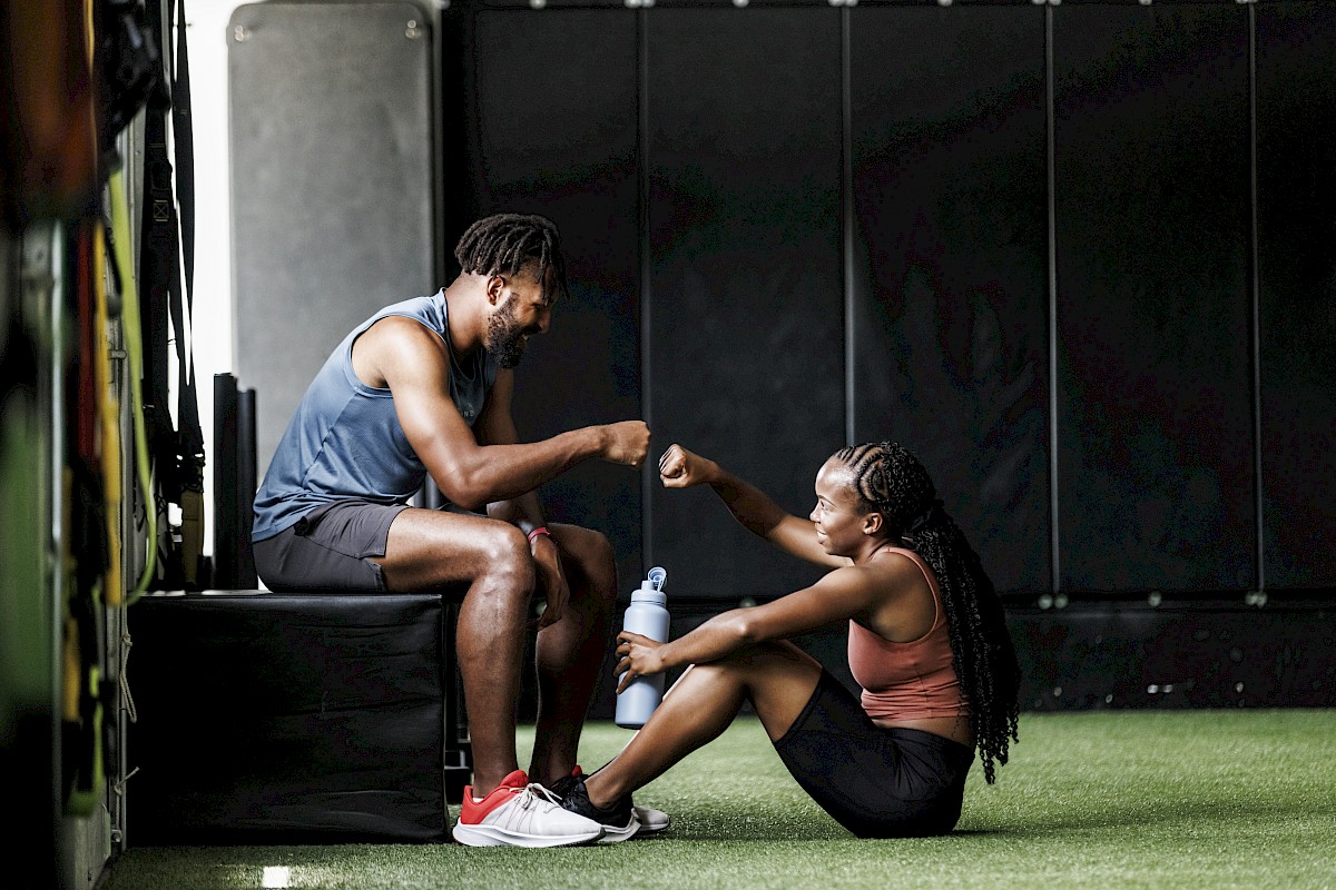 Two people in athletic gear are fist bumping in a gym, sitting on the floor and a bench, with one holding a water bottle.