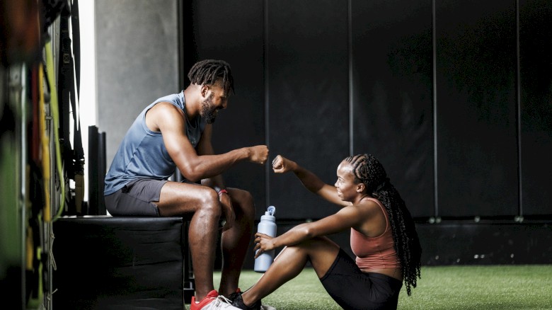 Two people in athletic gear are fist bumping in a gym, sitting on the floor and a bench, with one holding a water bottle.