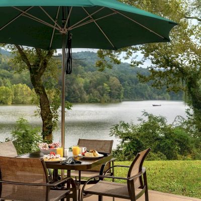 Outdoor dining setup under a green umbrella with a scenic view of a lake and surrounding trees. Four wicker chairs surround a table.