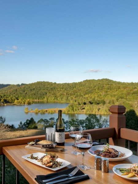 A table set with dishes, wine, and cutlery overlooks a scenic landscape with a lake and rolling hills under a clear blue sky, ending the sentence.