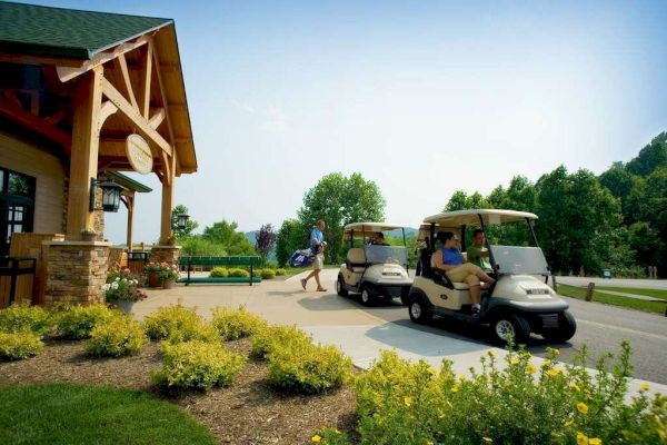 The image shows a golf course scene with two golf carts and a person walking while carrying a golf bag near a building surrounded by greenery.
