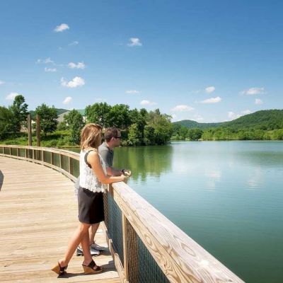 Two people stand on a wooden boardwalk overlooking a serene lake, surrounded by greenery and hills under a clear, blue sky, ending the sentence.