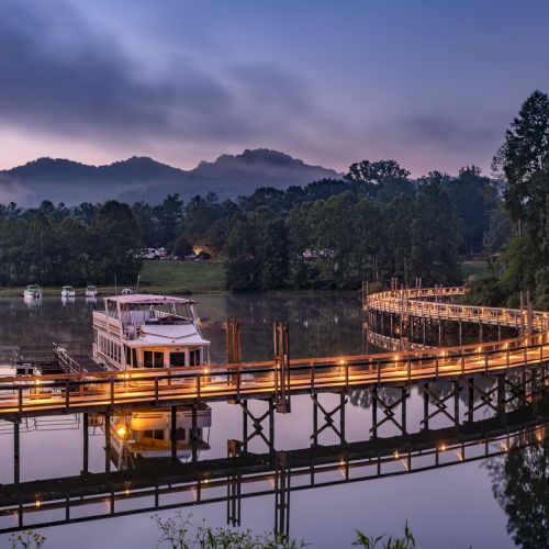 A tranquil scene featuring a lit wooden walkway over a lake, a boat docked at the end, surrounded by trees, and mountains in the background at dusk.