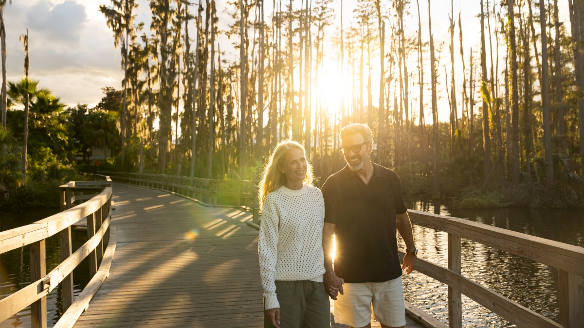 A couple walks hand-in-hand on a wooden bridge at sunset, surrounded by trees and water.