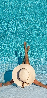 A person relaxing in a pool, wearing a wide-brimmed hat, with legs in the water and arms resting on the steps.