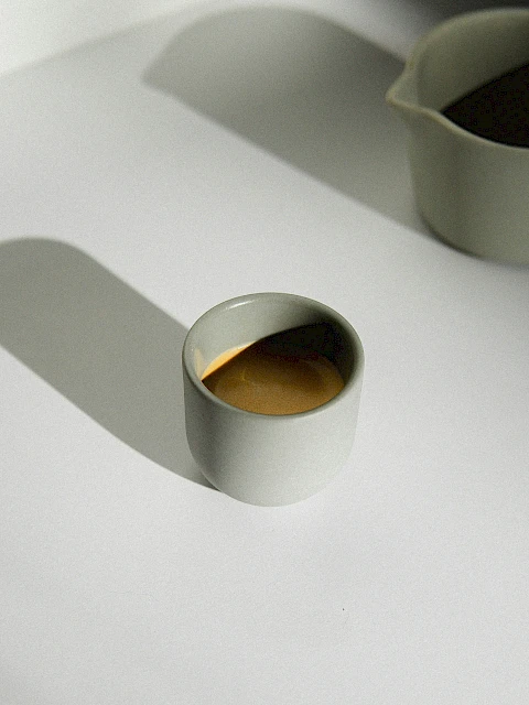 A small white cup filled with coffee sits on a white surface beside a matching jug casting shadows.