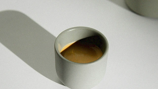 A small white cup filled with coffee sits on a white surface beside a matching jug casting shadows.