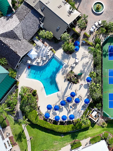 Aerial view of a recreational area with a pool, blue umbrellas, and multiple tennis courts surrounded by buildings and greenery.