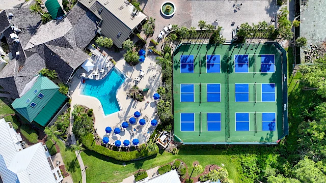 Aerial view of a recreational area with a pool, blue umbrellas, and multiple tennis courts surrounded by buildings and greenery.