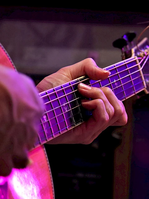 A person is playing an acoustic guitar, focusing on the hand fingering the strings with purple lighting.