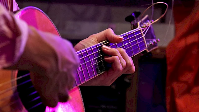 A person is playing an acoustic guitar, focusing on the hand fingering the strings with purple lighting.