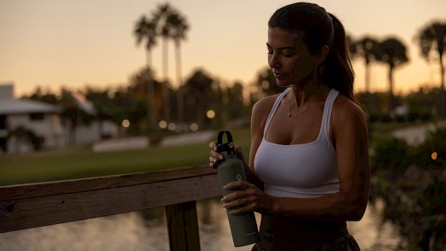 A person in workout attire holds a bottle on a bridge at sunset with palm trees in the background.