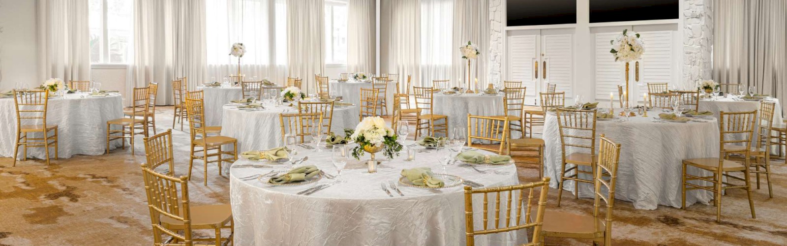 A beautifully decorated banquet hall with round tables, gold chairs, floral centerpieces, and gentle ambient lighting for a formal event.