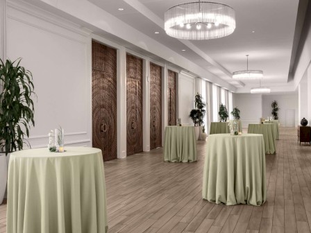 The image shows a well-lit hallway with round tables covered in green cloth, large potted plants, and wooden flooring.