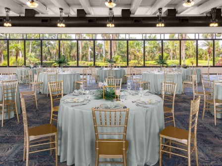 The image shows an elegantly set banquet hall with round tables, white tablecloths, and gold chairs, overlooking a garden through large windows.