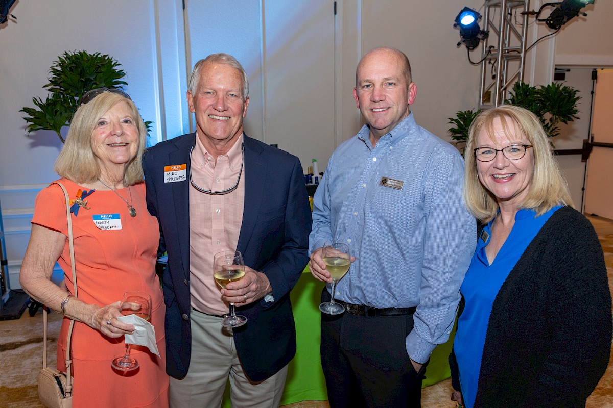 Four people are smiling at a social event, holding drinks and wearing name tags.