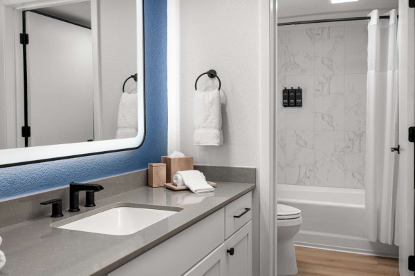 A modern bathroom with a large mirror, sink, black fixtures, towels, and a bathtub with a shower curtain in a marble-tiled setting.