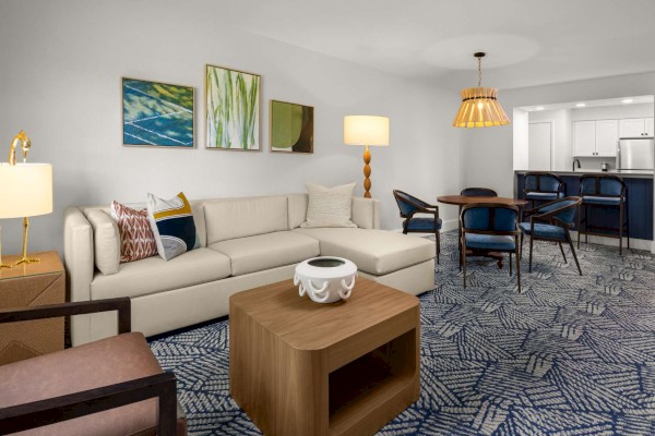 A modern living room with a beige sectional sofa, patterned rug, wooden coffee table, and adjacent dining area with blue chairs and hanging lamp.
