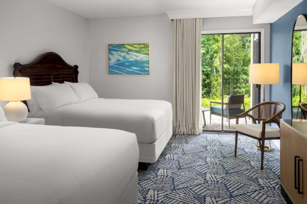 The image shows a hotel room with two beds, lamps, a painting, a chair, a TV, and a view of greenery through a glass door.
