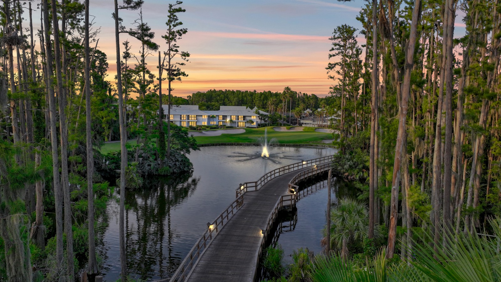 A serene lake with a wooden bridge, surrounded by tall trees, a building in the distance, and a fountain at sunset.