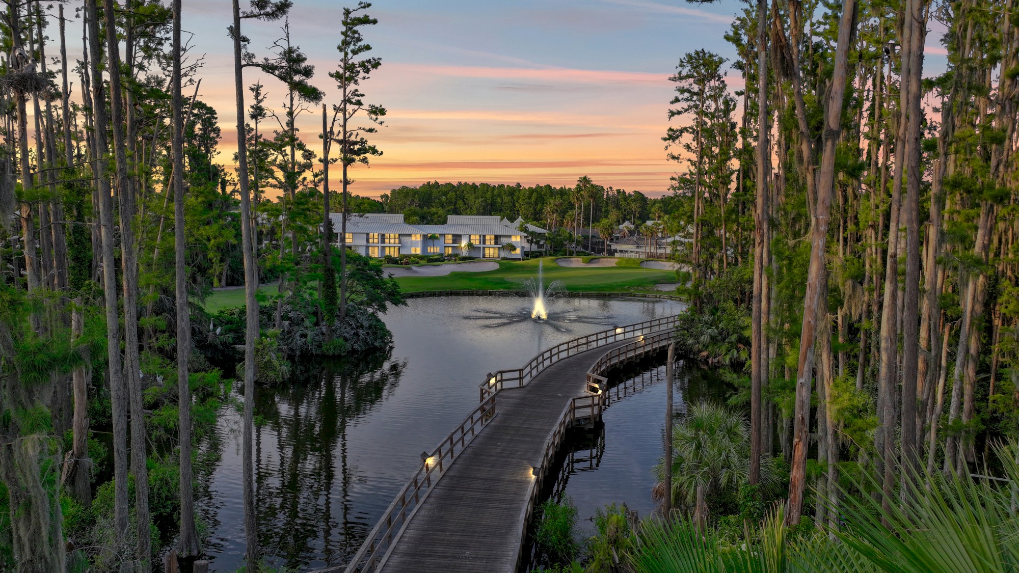 A serene lake with a wooden bridge, surrounded by tall trees, a building in the distance, and a fountain at sunset.