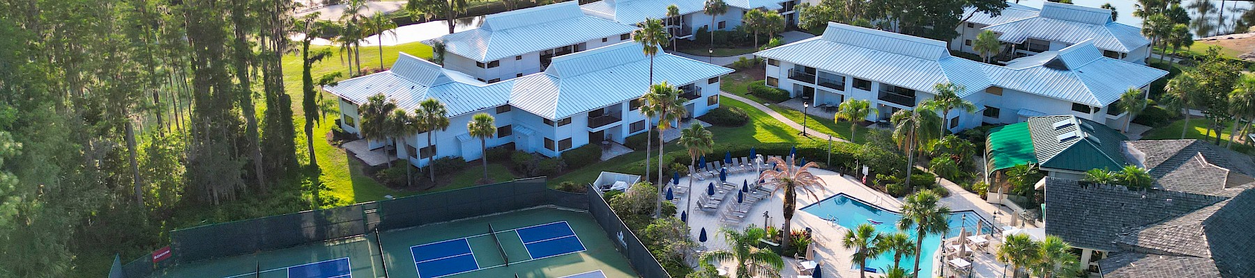 Aerial view of a resort with tennis courts, a pool, and several white-roofed buildings surrounded by trees and greenery.