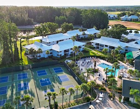 Aerial view of a resort with tennis courts, a pool, and several white-roofed buildings surrounded by trees and greenery.