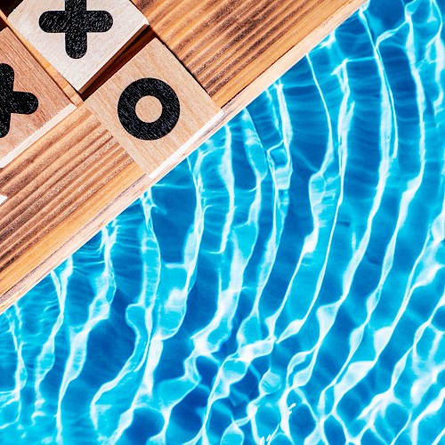 Tic-tac-toe game board with X and O pieces on a wooden surface, next to a pool with shimmering blue water.