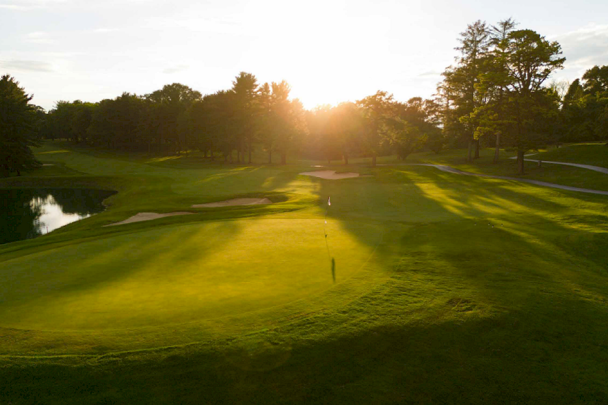 A golf course at sunset, with a green, water hazard, trees, and a flag on the putting green, creating a peaceful scene outdoors.