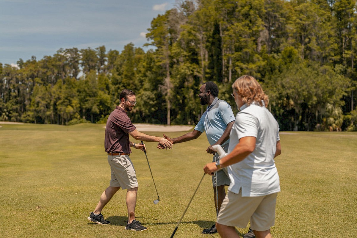 Three men shake hands on a golf course, engaging in friendly interaction during a sunny day.