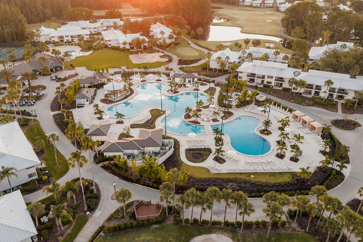 An aerial view of a resort with multiple pools, white buildings, palm trees, and a golf course in the background under warm sunlight.