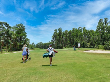 People walking on a golf course, carrying bags under a clear blue sky.