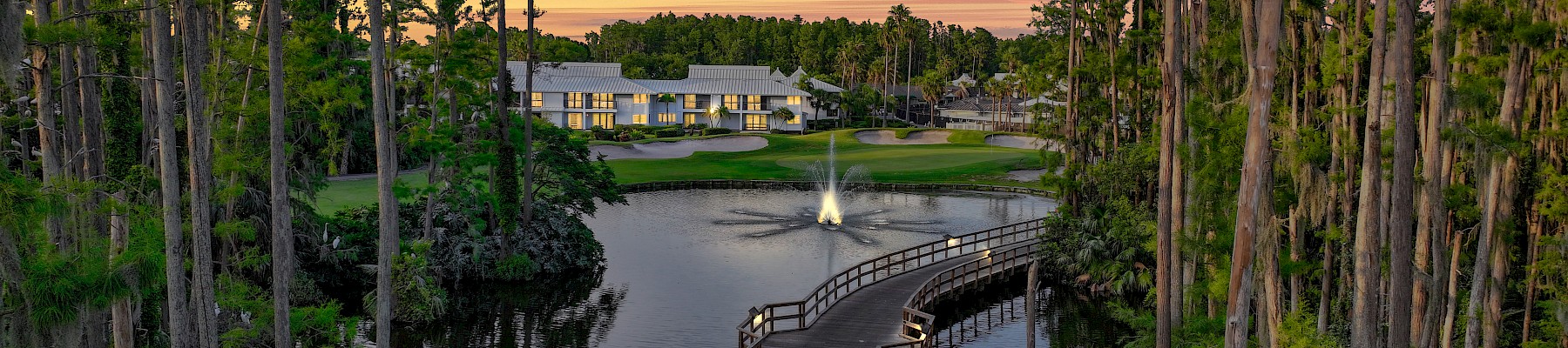 A serene landscape features a wood bridge over a pond, lined with tall trees, leading to a building in the background at sunset.
