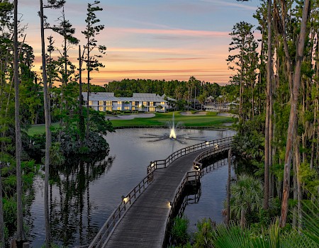 A serene landscape features a wood bridge over a pond, lined with tall trees, leading to a building in the background at sunset.