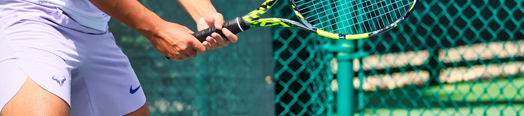 A person in white sportswear plays tennis on an outdoor court, holding a racket, near a green fence in sunny weather.