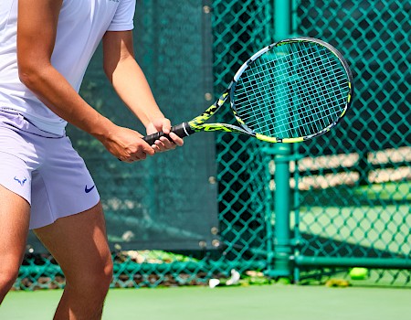 A person in white sportswear plays tennis on an outdoor court, holding a racket, near a green fence in sunny weather.