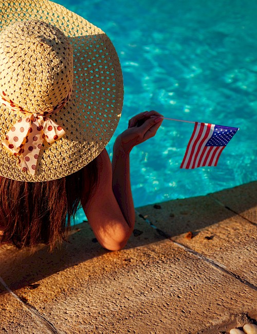 A person in a straw hat relaxes by a pool holding two small American flags.
