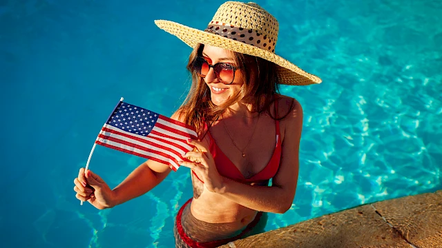 A woman in a pool smiles while holding a small American flag, wearing a sun hat and sunglasses.