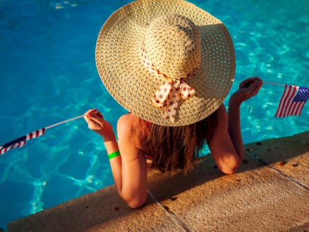 A person in a sun hat relaxes at a pool's edge, holding miniature American flags in both hands, surrounded by bright blue water.