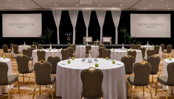 The image shows a conference room set up with round tables and chairs facing a stage with screens labeled 