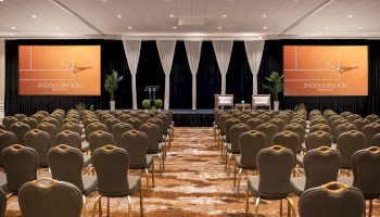 The image shows a conference room setup with rows of chairs facing a stage with two screens and plants beside it.