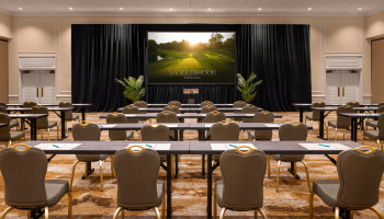 A conference room setup with rows of tables and chairs facing a stage with a large screen displaying 