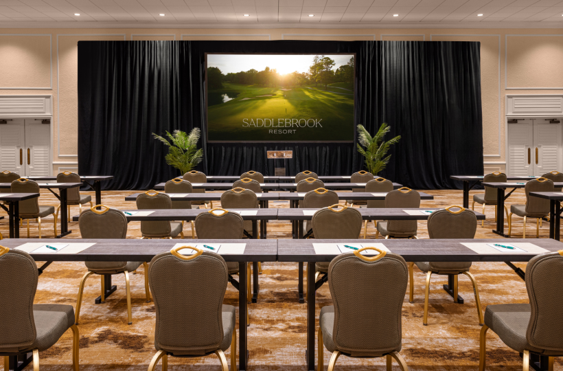 A conference room setup with rows of tables and chairs facing a stage with a large screen displaying 
