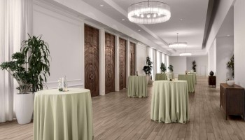 A modern banquet hall with round tables covered in green cloth, wooden flooring, large plants, and elegant ceiling lights.